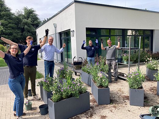 Gruppenbild nach erfolgreicher Bepflanzung von Blumenkübeln. Die bepflanzten Kübel sind zu sehen und fünf Personen, die fröhlich schauen. 
